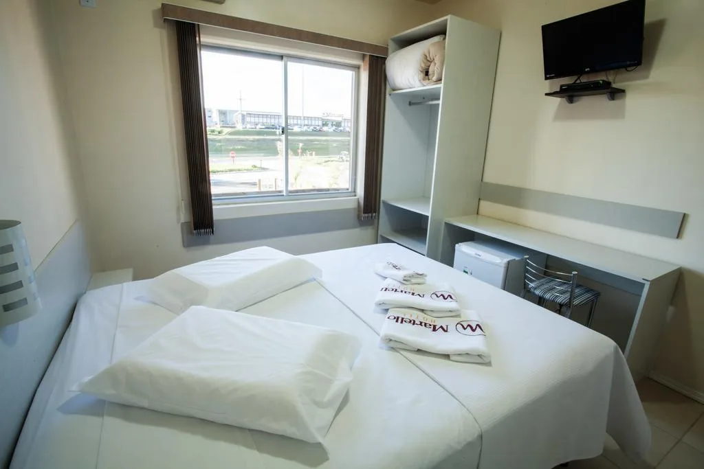 Quarto de hotel com cama de casal, duas toalhas brancas dobradas, TV na parede, janela com vista para cidade e prédios ao fundo