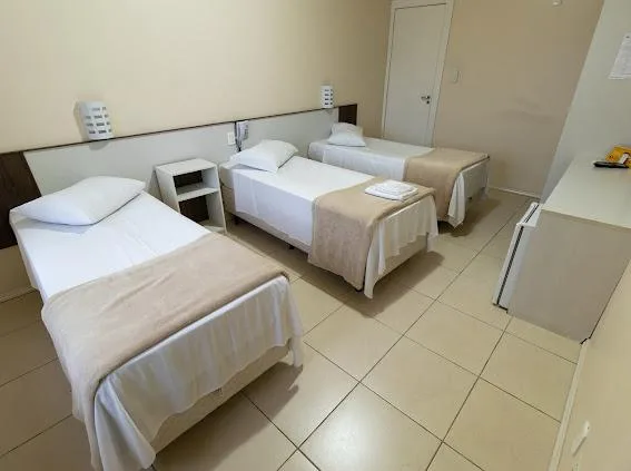 Quarto com três camas de solteiro com lençóis brancos, criado-mudo, piso em cerâmica clara e parede bege
