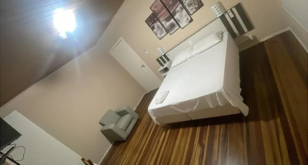 Quarto com cama branca, cadeira cinza, criado branco, quadros na parede, piso de madeira e luminária no teto