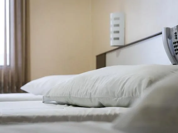 Dois travesseiros brancos sobre uma cama branca, com cabeceira de madeira ao fundo e parede bege com tomadas e cortinas