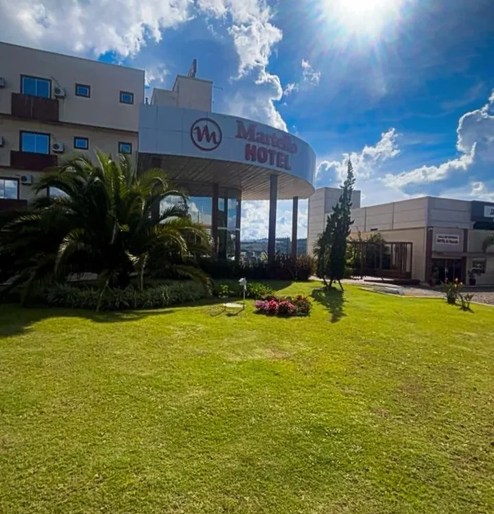Hotel com logo em fachada branca, gramado verde, palmeiras, flores coloridas, céu azul com nuvens e sol intenso