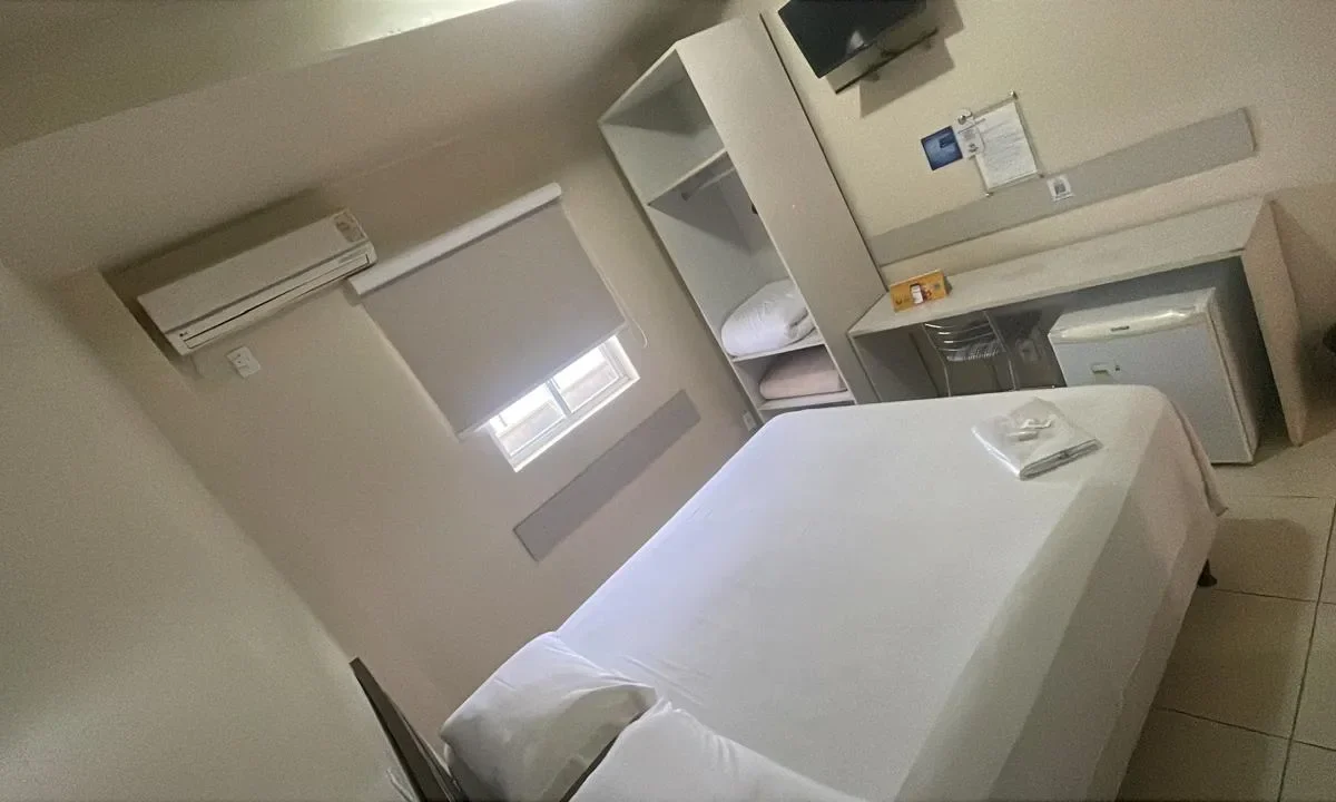 Quarto com cama de solteiro branca, ar-condicionado na parede, janela com cortina, prateleiras brancas, criado e porta à direita