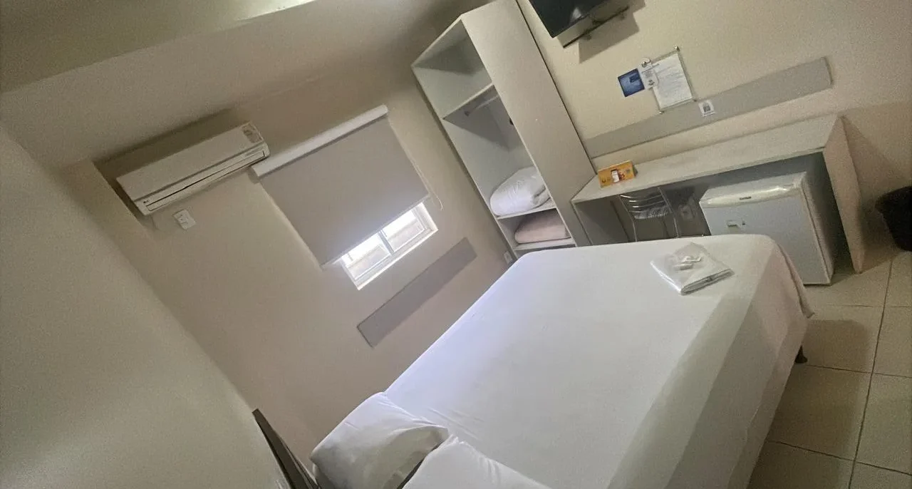 Quarto com cama de solteiro branca, ar-condicionado na parede, janela com cortina, prateleiras brancas, criado e porta à direita