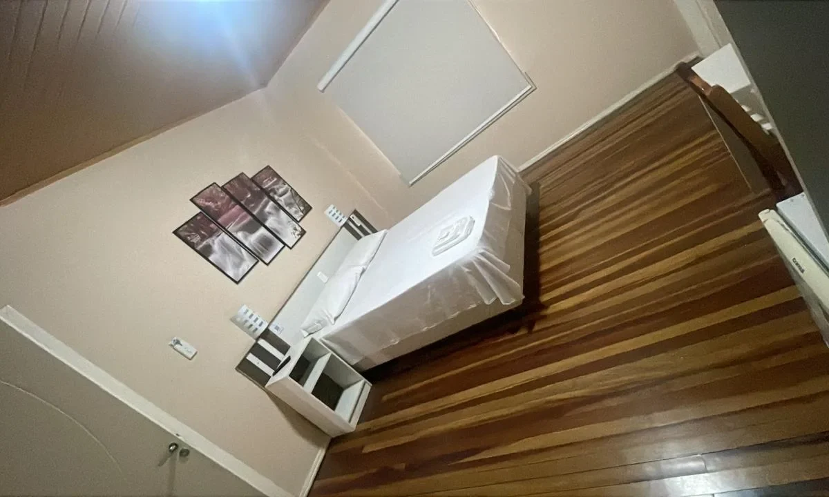 Quarto com cama branca, criado de cabeceira de madeira, quadros na parede bege e piso de madeira clara