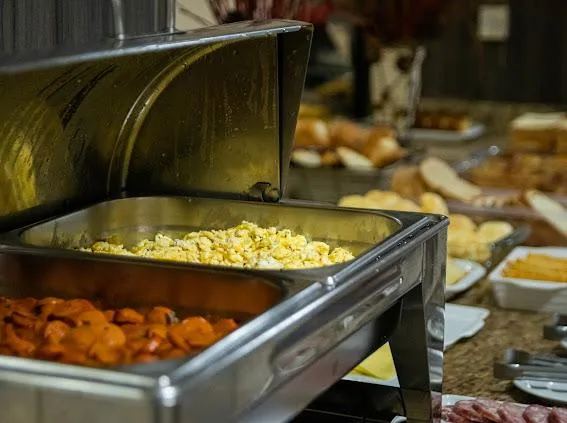 Bandejas metálicas com alimentos sendo servidos, incluindo milho cozido em amarelo e cenoura em laranja, em ambiente de cozinha