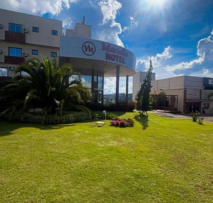 Hotel com logo em fachada branca, gramado verde, palmeiras, flores coloridas, céu azul com nuvens e sol intenso