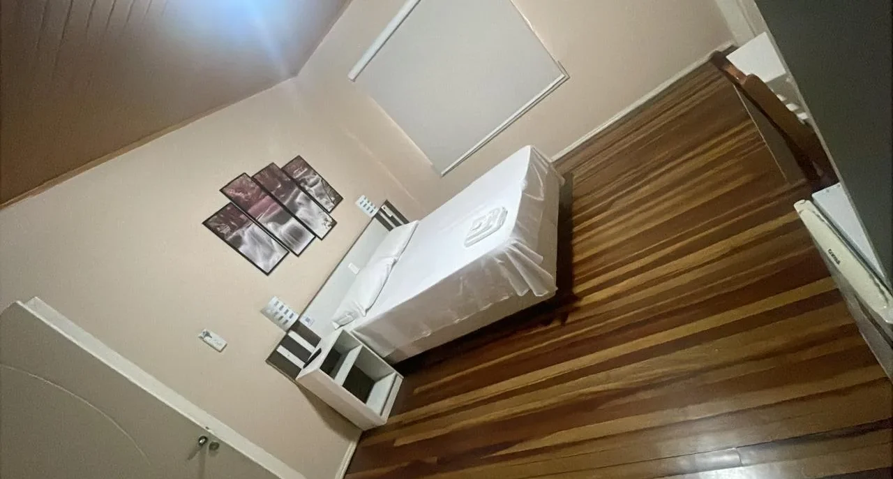 Quarto com cama branca, criado de cabeceira de madeira, quadros na parede bege e piso de madeira clara