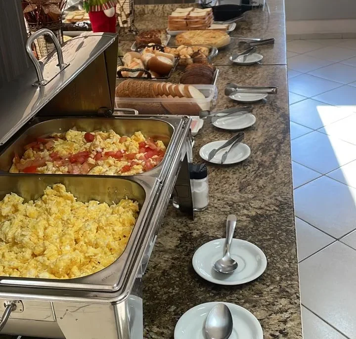 Balcão com bandejas de comida, pratos brancos com colheres, pães, decoração floral vermelha e branca, relógio de parede e janelas ao fundo