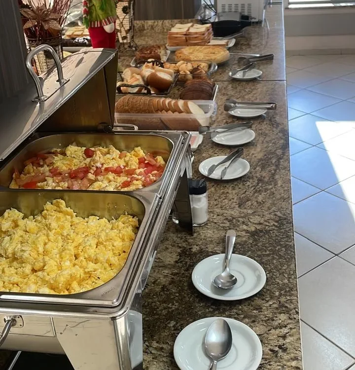 Balcão com bandejas de comida, pratos brancos com colheres, pães, decoração floral vermelha e branca, relógio de parede e janelas ao fundo