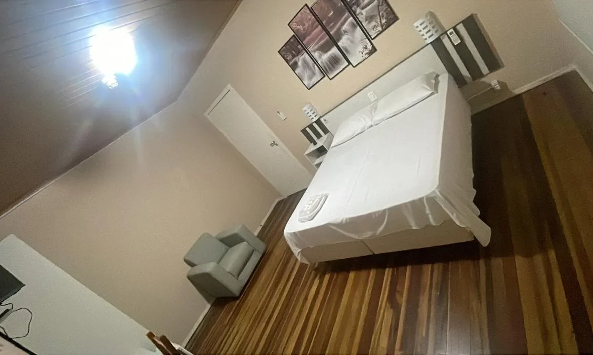 Quarto com cama branca, cadeira cinza, criado branco, quadros na parede, piso de madeira e luminária no teto
