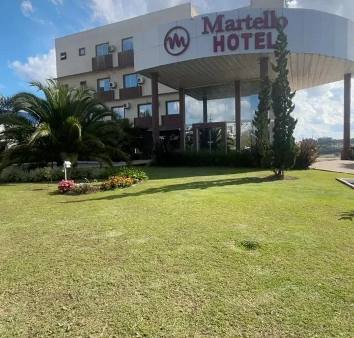 Hotel Martello com arquitetura moderna, fachada branca e vermelha, amplo gramado verde, palmeiras e cipreste, céu azul com nuvens brancas