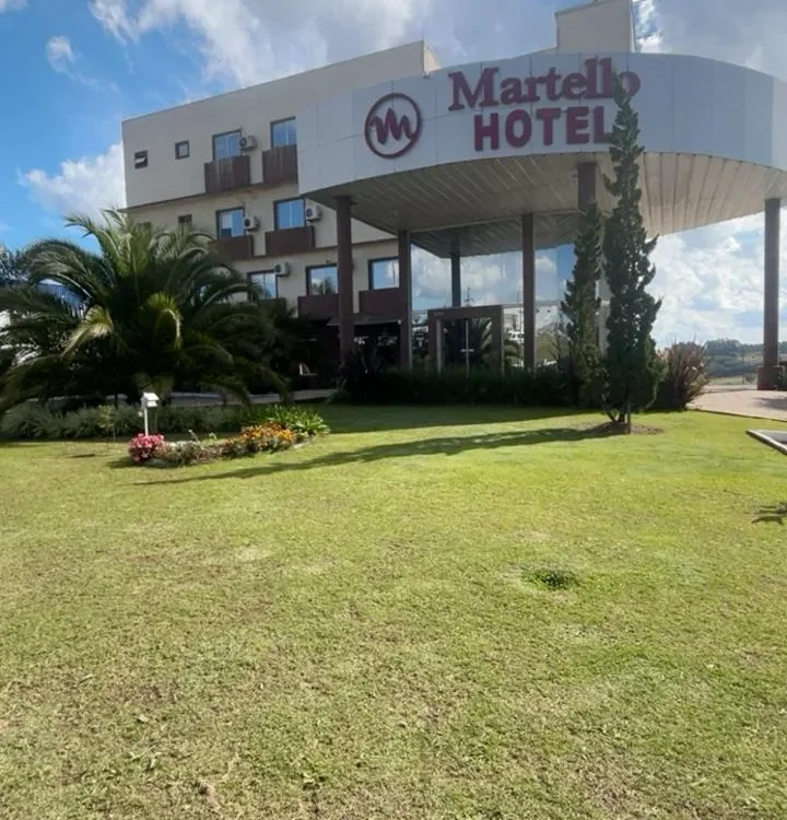 Hotel Martello com arquitetura moderna, fachada branca e vermelha, amplo gramado verde, palmeiras e cipreste, céu azul com nuvens brancas